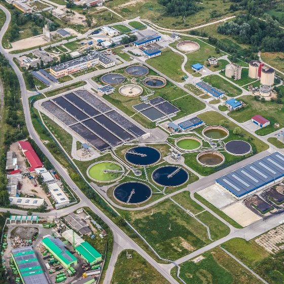 Aerial photo of wastewater facility in a field with several cylindrical and rectangular treatment tanks and buildings all connected by paved roads and paths.