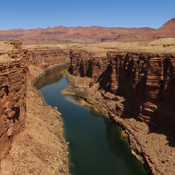 A dark river with a still surface flows at the base of tall rocky banks in an arid landscape with a reddish mountain range in the background under clear blue skies.