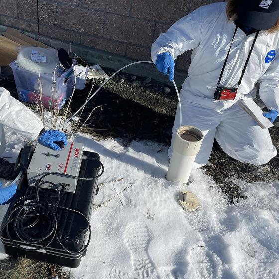 Two people in white protective suits and blue gloves kneel with their testing equipment in front of a brick wall and insert a plastic tube in a vertical white pipe emerging from snow-covered ground.