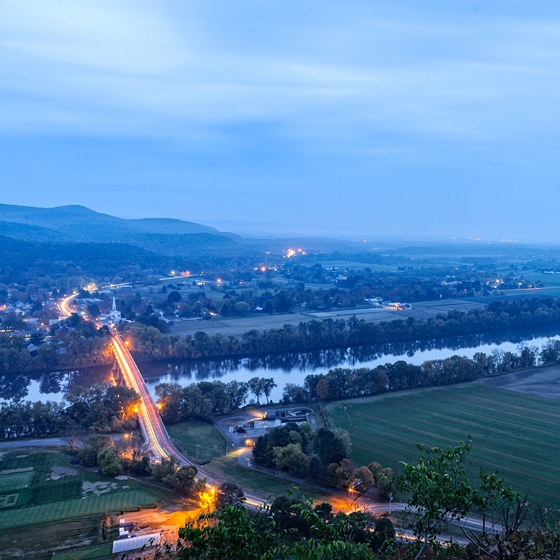 An aerial shot of a landscape in low light, bisected horizontally by a tree-lined river and vertically by a paved road dotted with orange streetlights and running through a town at the base of a mountain.