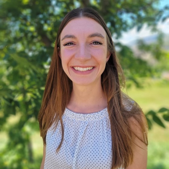 Natalie Brunel, Junior Environmental Scientist, stands outside wearing business casual attire in front of a leafy green background.