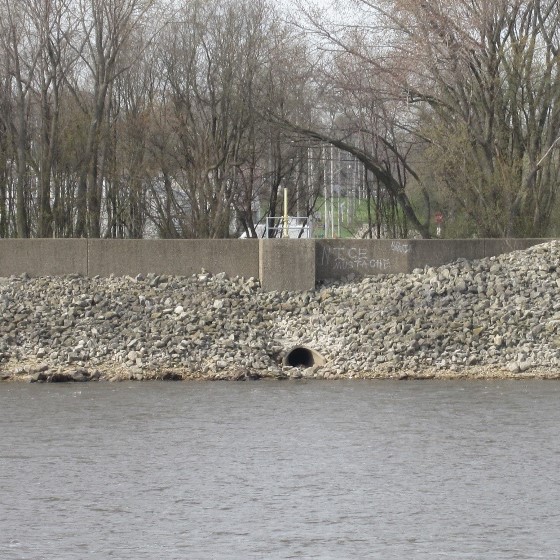 Image of wastewater outfall adjacent to a waterbody lined by a rocky embankment with bare trees in the background.